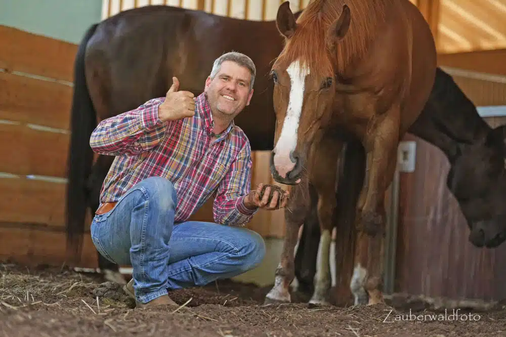 Bernd Hackl Jeans hockt in einer Stallgasse zwischen zwei Pferden und zeigt den Daumen nach oben. Das braune Pferd mit weißer Blesse steht nah an der Person und blickt interessiert auf die Hand. Im Hintergrund sind Holzstallwände und ein weiteres Pferd zu sehen.