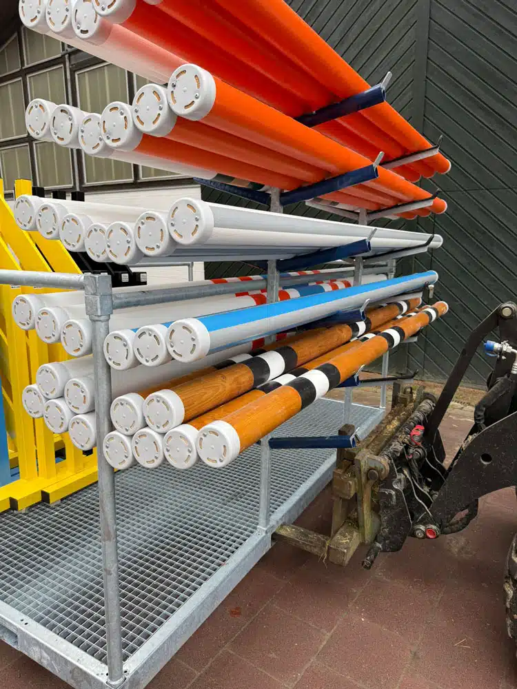 Detaillierte Nahaufnahme eines Metallgestells mit mehreren farbig lackierten Stangen, die für Hindernisse im Reitsport verwendet werden. Zu erkennen sind orange, weiße, blaue und braune Stangen, die horizontal gestapelt sind. Links sind gelbe und blaue Hindernis-Elemente aufrecht einsortiert. Im Hintergrund befindet sich ein Teil eines landwirtschaftlichen Fahrzeugs sowie die dunkle Fassade einer Halle.