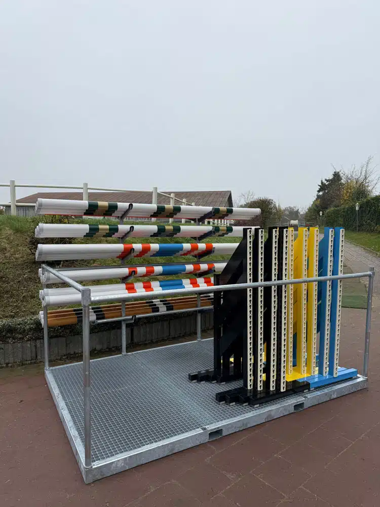 Ein Metallgestell im Freien dient als Aufbewahrungssystem für Reitsport-Equipment. Darin sind farbig bemalte Stangen horizontal gestapelt und mehrere Hindernissständer mit höhenverstellbaren Löchern senkrecht sortiert. Der Untergrund besteht aus einer geriffelten Metallplatte, im Hintergrund sind ein Zaun, Rasenfläche und einige Gebäude zu sehen.
