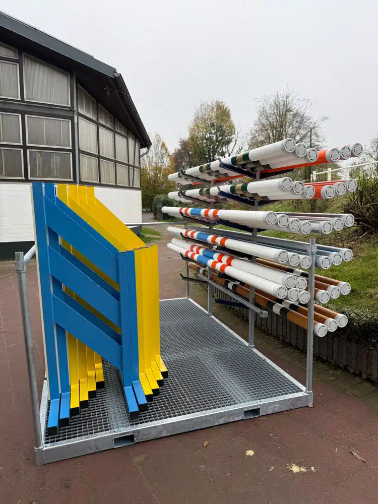 Auf dem Bild ist ein stabiles, rechteckiges Metallgestell im Außenbereich zu sehen, das zur Aufbewahrung von Reitsport-Hindernissen dient. Rechts sind zahlreiche weißgrundierte, farbig gestreifte Stangen horizontal übereinandergestapelt. Links stehen blaue und gelbe Hindernis-Elemente vertikal einsortiert. Der Boden des Gestells besteht aus einer geriffelten Metallplatte, im Hintergrund befinden sich eine Halle, Rasenfläche, Sträucher und Bäume.