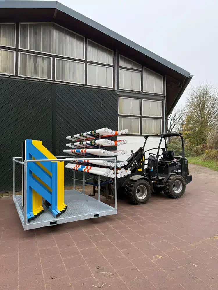 Ein Kompakttraktor transportiert ein Metallgestell, das zur Aufbewahrung von bunten Hindernisstangen und blauen sowie gelben Hindernis-Elementen für den Reitsport dient. Im Hintergrund steht eine Reithalle mit dunkler Holzfassade und großen Fenstern; die Szene spielt auf einer gepflasterten Fläche im Außenbereich.