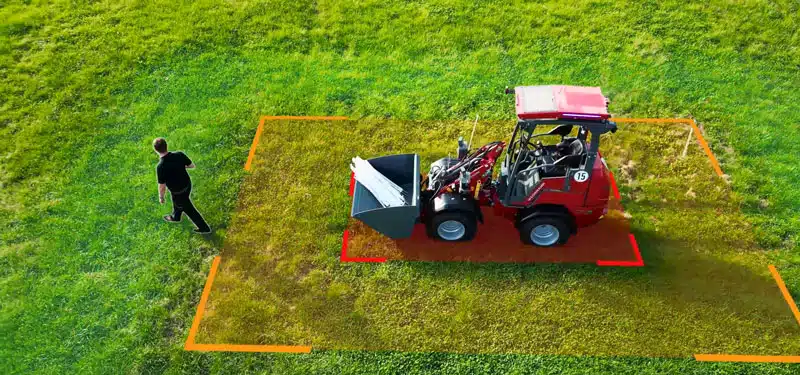 Auf einer grünen Wiese steht ein kompakter roter Weidemann Radlader mit Frontschaufel, neben dem eine Person geht. Rund um das Fahrzeug ist ein leuchtend orangefarbenes Rechteck auf dem Gras markiert, das vermutlich einen Sicherheitsabstand kennzeichnet. Die Szene ist aus der Vogelperspektive aufgenommen.